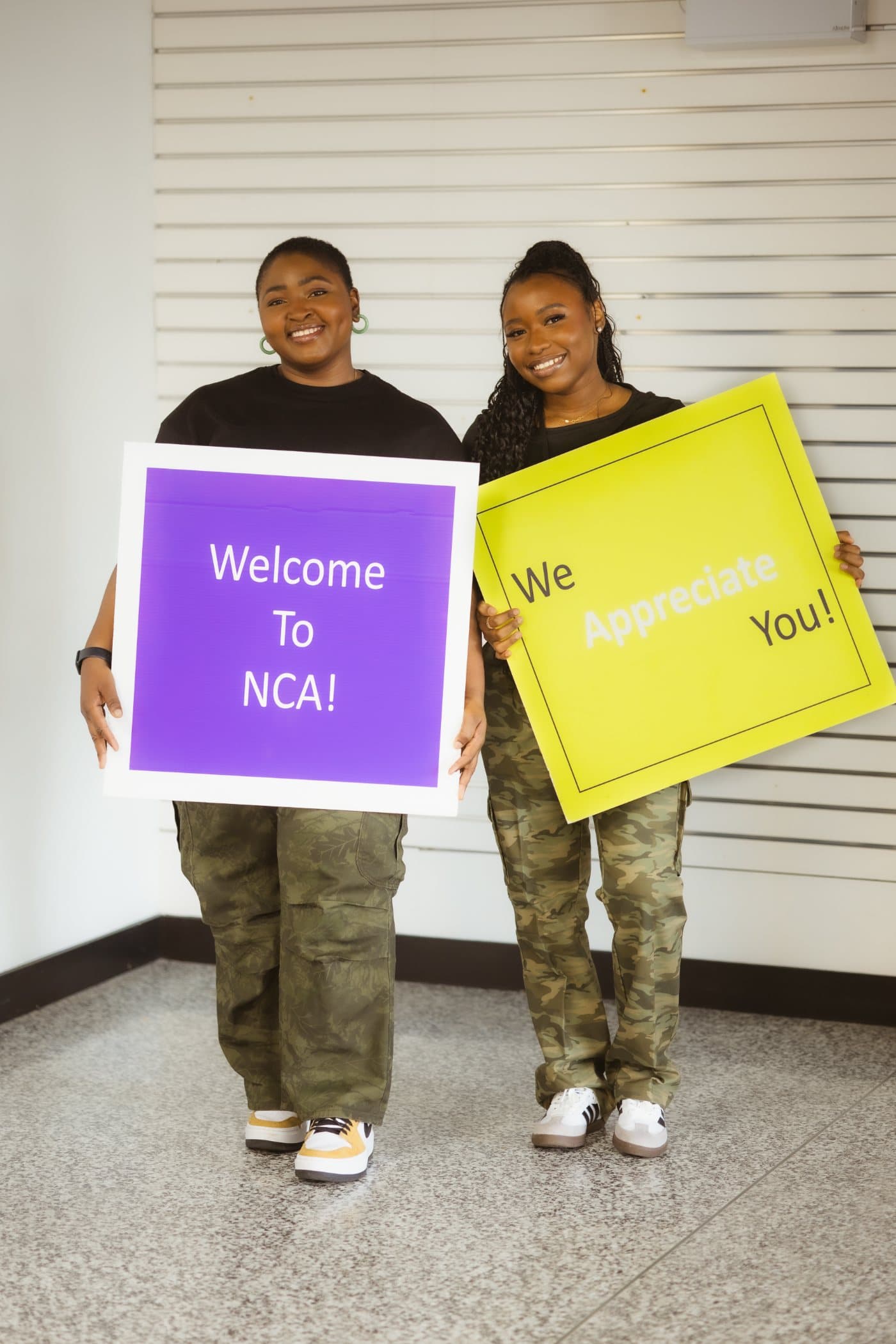 Deux bénévoles de NCA souriants tenant des pancartes où l’on lit Welcome to NCA et We appreciate you